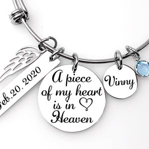 May include: A silver bracelet with a charm that reads "A piece of my heart is in Heaven" and a heart symbol. The bracelet also has a charm with the text "Vinny" and a small blue crystal charm. A separate charm has the text "Feb. 20, 2020" and a wing design.