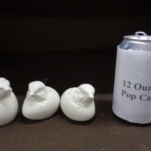 May include: Three white ceramic bird figurines, each depicting a bird emerging from a cracked egg shell. The figurines are arranged in a row on a brown surface. A 12-ounce pop can is shown for size comparison.
