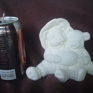 May include: White ceramic figurine of two teddy bears hugging. The bear on the left is wearing a hat. The figurine is approximately 6 inches tall.
