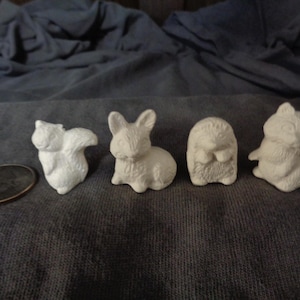 May include: Four small, white ceramic figurines of woodland animals. The figurines are of a squirrel, a rabbit, a hedgehog, and a raccoon.