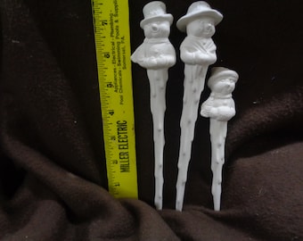 May include: Three white ceramic snowman icicle ornaments. The snowmen are wearing hats and have a whimsical look. The icicles are approximately 18 cm long.