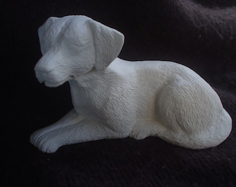 May include: White ceramic sculpture of a dog lying down with its head resting on its paws. The sculpture is detailed with fur texture.