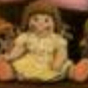 May include: A small porcelain doll wearing a yellow dress and with brown hair. The doll is sitting on a wooden surface.
