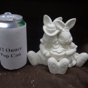 May include: White ceramic figurine of two bunnies holding a heart-shaped object. The bunnies are wearing hats and the figurine is about the size of a 12-ounce pop can.