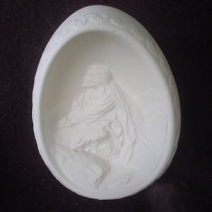 May include: White ceramic egg-shaped ornament with a raised relief image of a person inside. The person is wearing a robe and has a halo above their head.