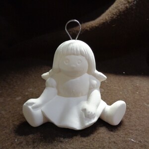 May include: White ceramic ornament of a girl doll sitting with her legs crossed. The doll has a simple dress and a wire hanger on top of her head.