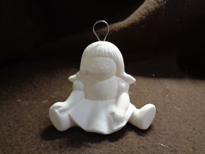 May include: White ceramic ornament of a girl doll sitting with her legs crossed. The doll has a simple dress and a wire hanger on top of her head.