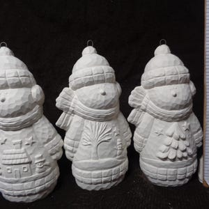 May include: Three unpainted ceramic snowman ornaments. Each snowman wears a hat and scarf. One has a house design, another a tree, and the last a Christmas tree. Each ornament has a metal loop for hanging.