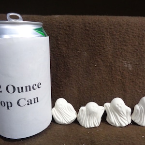 May include: A silver pop can with a white paper sleeve that reads "12 Ounce Pop Can." Four small, white ghost figurines are lined up on a brown surface. The ghosts have simple, smiling faces and draped sheet-like bodies.