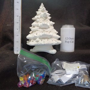 May include: A white ceramic Christmas tree with a textured surface, next to a ruler, a 12-ounce pop can, a bag of colorful lights, and a bag of electrical components. The tree is approximately 7 inches tall.