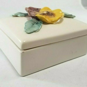 May include: A white ceramic box with a raised floral design. The flower is yellow with brown and green accents. The box has a hinged lid.