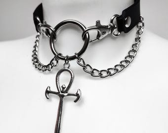 May include: A black choker necklace featuring a silver-toned metal ring at the center, connected to two silver-toned metal chains. A silver-toned ankh cross pendant hangs from the central ring. The choker is secured with silver-toned metal hardware.