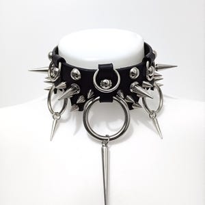 May include: A black leather choker with silver spikes and rings. The choker has a large ring in the center with a long silver spike hanging from it.
