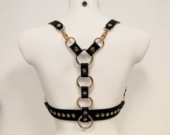 Pode incluir: Arnês de couro preto com anéis e tachas de metal dourado. O arnês tem um decote em V e correias ajustáveis.