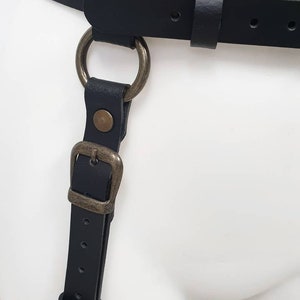 Puede incluir: Arn&eacute;s de cuero negro con un gran anillo y hebilla de metal. El arn&eacute;s tiene correas que van alrededor de la cintura y los muslos.