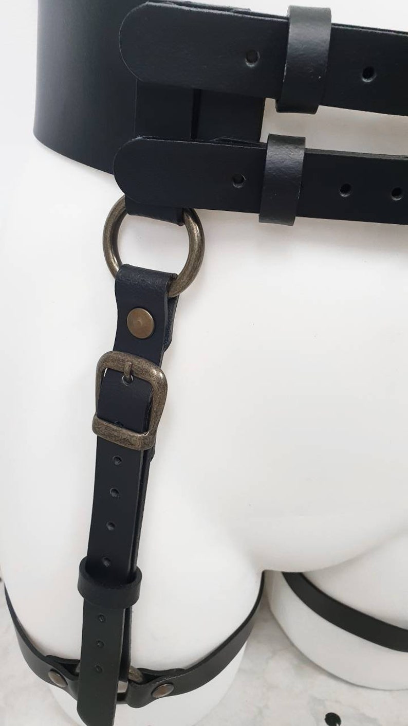 Puede incluir: Arn&eacute;s de cuero negro con un gran anillo y hebilla de metal. El arn&eacute;s tiene correas que van alrededor de la cintura y los muslos.