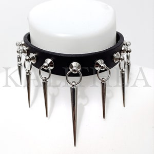 Puede incluir: Choker de cuero negro con púas y anillos plateados. El choker tiene una fila de tachuelas plateadas y una fila de anillos plateados con púas que cuelgan.