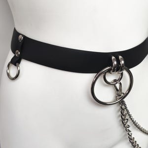 May include: A black belt featuring silver-toned metal hardware, including a single side ring attached with two rivets and a large double-ring centerpiece. A silver-toned metal chain hangs from the central rings, adding a layered metallic detail.