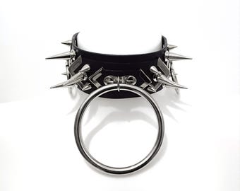 Puede incluir: Un collar de cuero negro con pinchos plateados y un anillo plateado grande. El collar está decorado con formas geométricas.