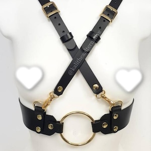 May include: Black leather body harness with gold-colored hardware, including a large ring and adjustable straps. The harness features a crisscross design across the chest and is secured with buckles and snap closures. The text "@kalithealternative" is visible on the straps.