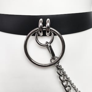 May include: A black belt featuring a large silver-toned metal ring attached to two smaller silver-toned rings. A silver-toned metal lobster clasp connects a double-layered silver-toned chain to the large ring, creating a dangling hardware detail.