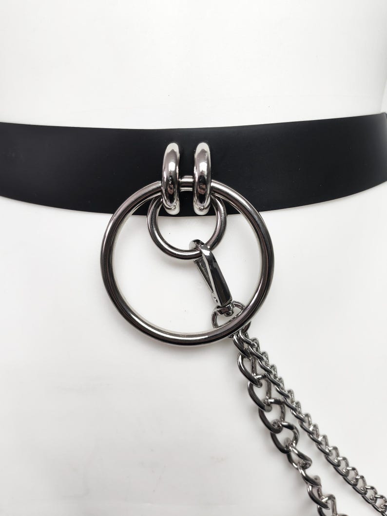 May include: A black belt featuring a large silver-toned metal ring attached to two smaller silver-toned rings. A silver-toned metal lobster clasp connects a double-layered silver-toned chain to the large ring, creating a dangling hardware detail.
