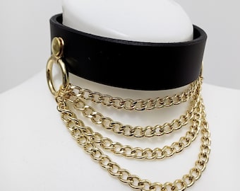May include: Black leather choker with a gold metal ring and multiple gold chains hanging from it.