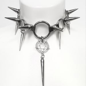 以下が含まれることがあります： 複数のスパイクとペンタグラムのペンダントが付いたシルバーのチョーカーネックレス。ネックレスは白いマネキンにかけられています。