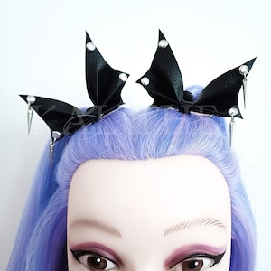 Puede incluir: Pinzas para el pelo de cuero negro en forma de alas de murciélago con detalles plateados. Las pinzas se llevan en una modelo con el pelo morado.
