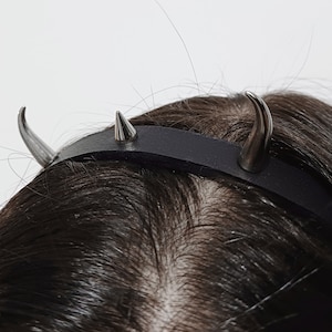 Könnte beinhalten: Ein schwarzes Leder Stirnband mit zwei silbernen Spikes. Das Stirnband wird auf dem Kopf einer Person getragen.