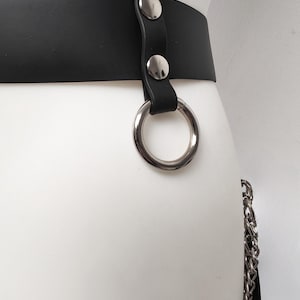 May include: A black belt featuring a smooth matte finish and a silver-toned metal O-ring attached with a vertical strap secured by two polished silver-toned rivets. A silver-toned metal chain hangs from the side of the belt.
