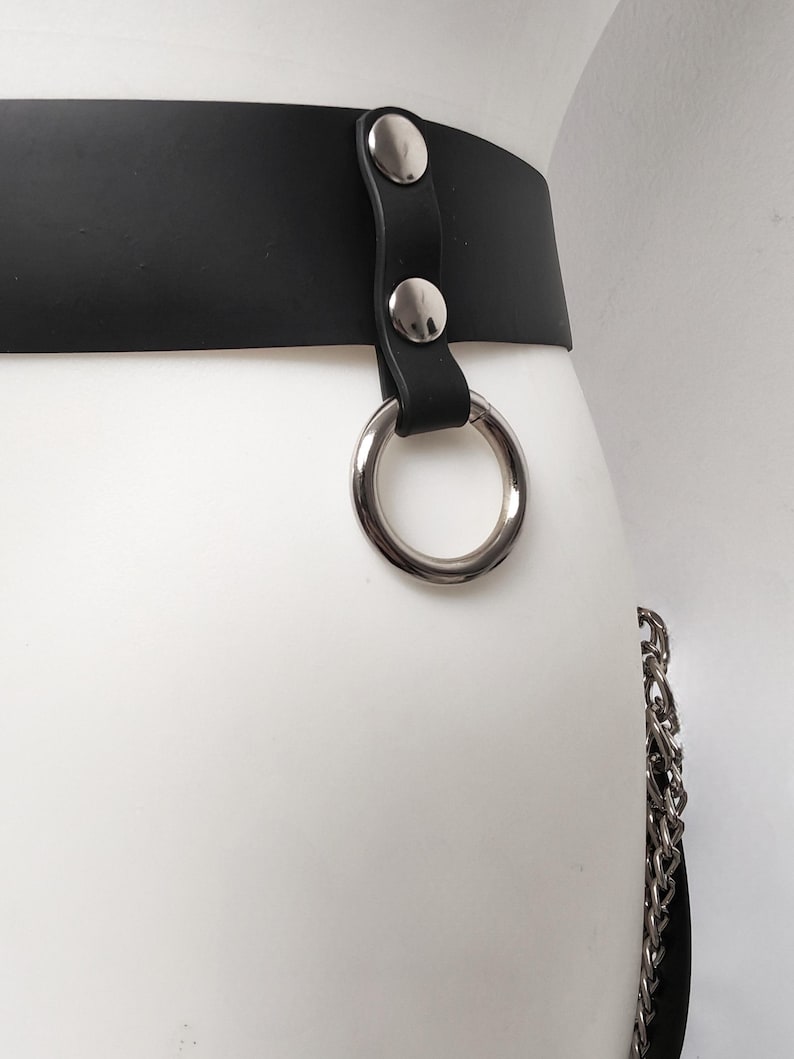 May include: A black belt featuring a smooth matte finish and a silver-toned metal O-ring attached with a vertical strap secured by two polished silver-toned rivets. A silver-toned metal chain hangs from the side of the belt.