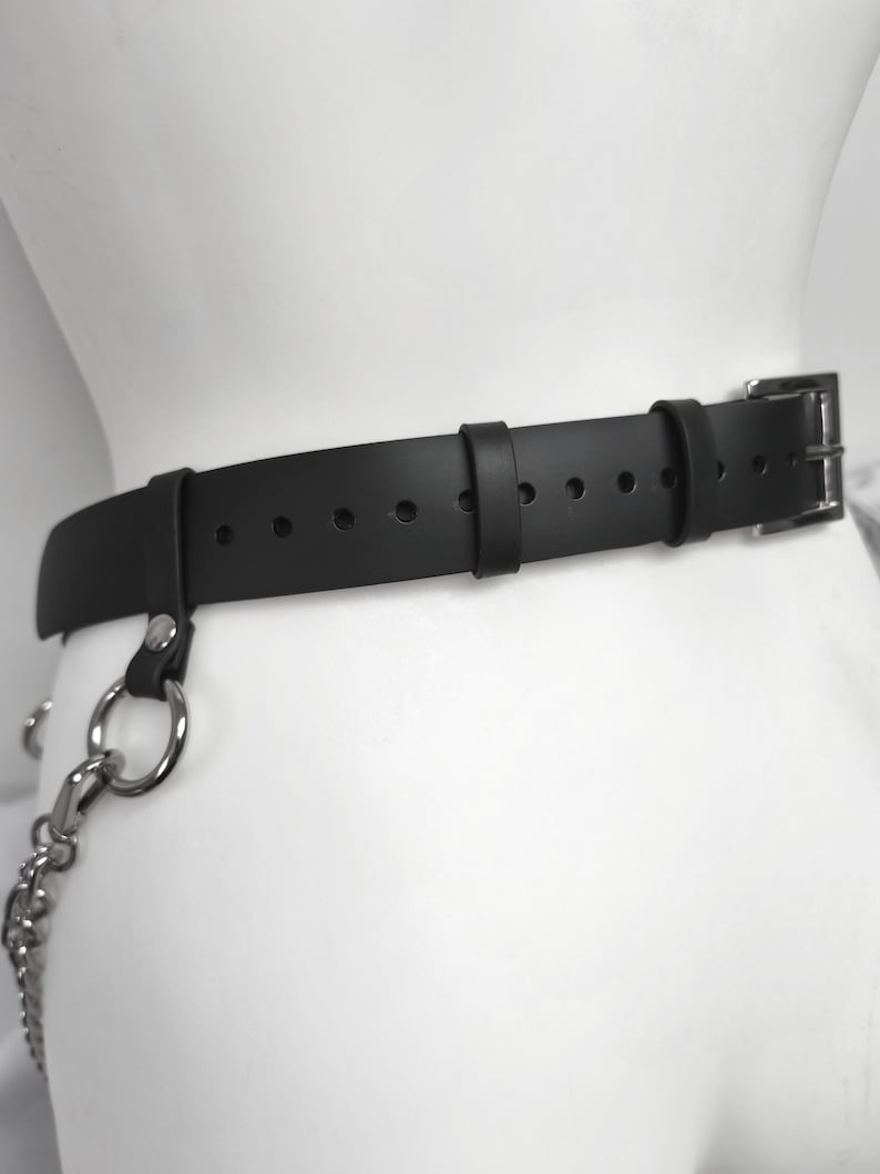 May include: A black belt featuring a silver-toned metal buckle and a series of adjustment holes. The belt includes two fixed loops and a silver-toned metal ring attached to a side strap, which connects to a dangling silver-toned metal chain.