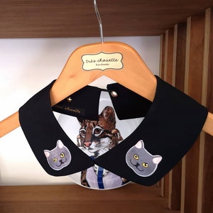 Könnte beinhalten: Schwarzer abnehmbarer Kragen mit zwei gestickten grauen Katzenköpfen. Der Kragen befindet sich auf einem Holzbügel mit einem Etikett, auf dem "très chouette handmade" steht.