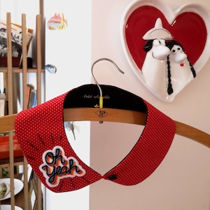 Puede incluir: Collar de tela roja con lunares blancos y un parche colorido "Oh Yeah". El collar está en una percha de madera.