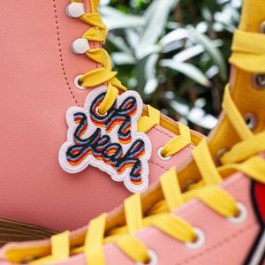 Peut inclure: Des baskets roses à lacets jaunes et des patchs colorés. Un patch dit "Yeah!" et l'autre est un cœur rouge avec un bâton de sucette blanc.