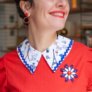 Könnte beinhalten: Eine rote Bluse mit einem abnehmbaren Kragen mit weißem und blauem Muster. Der Kragen zeigt ein Pariser Thema mit Illustrationen des Eiffelturms, Weingläsern und anderen Pariser Sehenswürdigkeiten. Eine rot-weiß-blaue Blumenbrosche ist an der Bluse befestigt.