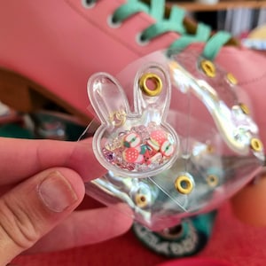 May include: A clear, bunny-shaped charm filled with colorful beads and fruit-shaped confetti. The charm is held in front of a pink roller skate with teal laces. The charm has gold-colored eyelets.