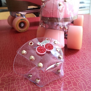 May include: Pink roller skates with clear plastic toe guards decorated with rhinestones and a cherry design. The toe guards are secured with gold-colored rivets.