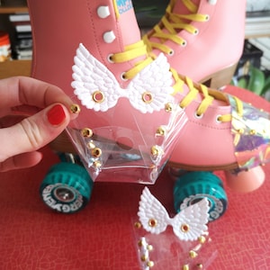 Puede incluir: Patines rosas con cordones amarillos y alas de ángel blancas fijadas en la parte delantera. Los patines tienen protectores de dedos de plástico transparente con tachuelas doradas.