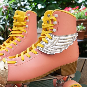 May include: A pair of pink roller skates with yellow laces and gold accents. The skates have a silver wing design on the side.