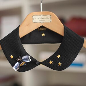 May include: A black fabric collar with gold star embellishments and a bee-shaped brooch. The collar is hanging on a wooden hanger with a label that reads "Très chouette".