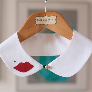 Puede incluir: Un cuello de fieltro blanco con una aplicación de labios rojos y un pequeño punto negro. El cuello cuelga de una percha de madera con el texto "très chouette handmade". Se abrocha con un botón dorado.