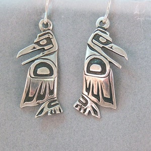 Puede incluir: Un par de pendientes de plata con un diseño estilizado de pájaro. Cada pendiente tiene un patrón grabado detallado y un gancho para orejas perforadas. Los pendientes se muestran sobre un fondo gris.
