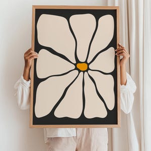 May include: A framed art print featuring a large, stylized flower. The petals are a creamy beige against a black background, with a bright yellow center. The frame is a light wood color, adding a natural touch to the modern floral design.