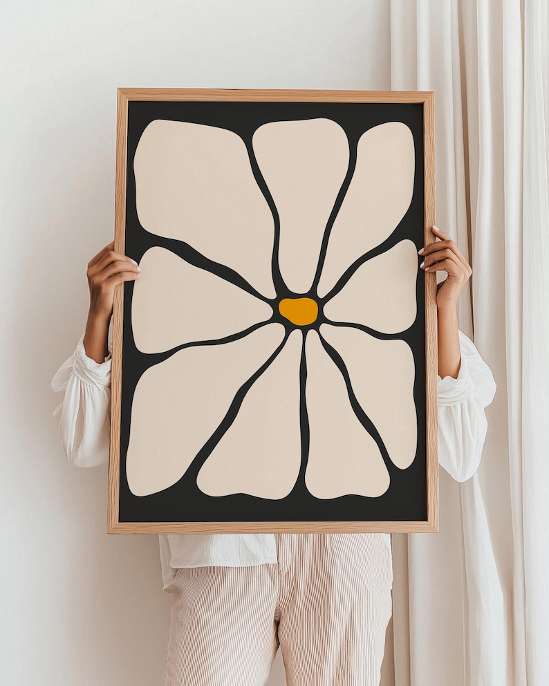 May include: A framed art print featuring a large, stylized flower. The petals are a creamy beige against a black background, with a bright yellow center. The frame is a light wood color, adding a natural touch to the modern floral design.