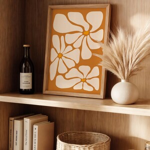 May include: A framed print with a minimalist abstract design of white flowers on a mustard yellow background. The print is displayed on a wooden shelf with a vase of dried pampas grass, books, and a wicker basket.