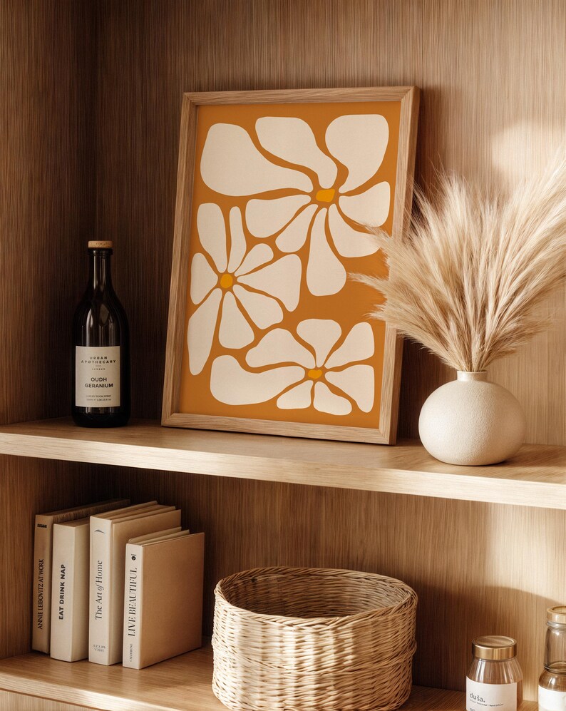 May include: A framed print with a minimalist abstract design of white flowers on a mustard yellow background. The print is displayed on a wooden shelf with a vase of dried pampas grass, books, and a wicker basket.
