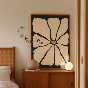 May include: A large framed art print featuring a stylized daisy with cream-colored petals and a yellow center against a black background. The artwork is displayed above a wooden cabinet with a vase, books, and a small lamp.