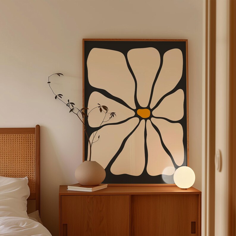 May include: A large framed art print featuring a stylized daisy with cream-colored petals and a yellow center against a black background. The artwork is displayed above a wooden cabinet with a vase, books, and a small lamp.